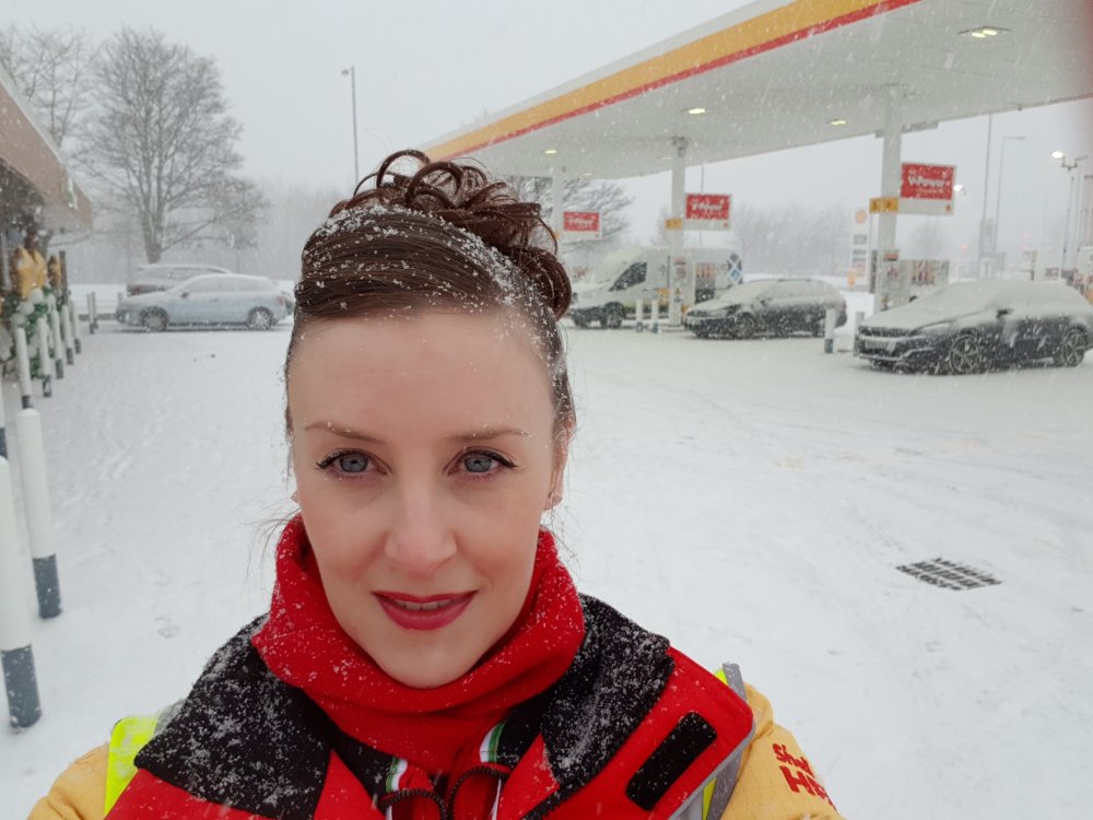 Nicola taking a selfie in the snow wearing a Shell high-vis uniform, standing in front of a Shell petrol station during extreme winter weather.
